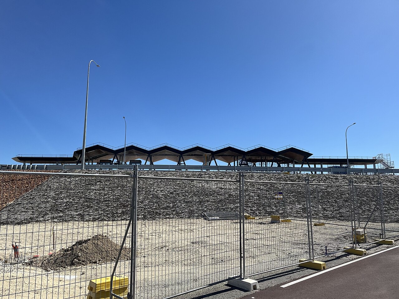 A construction site with a long roof structure behind temporary fencing.